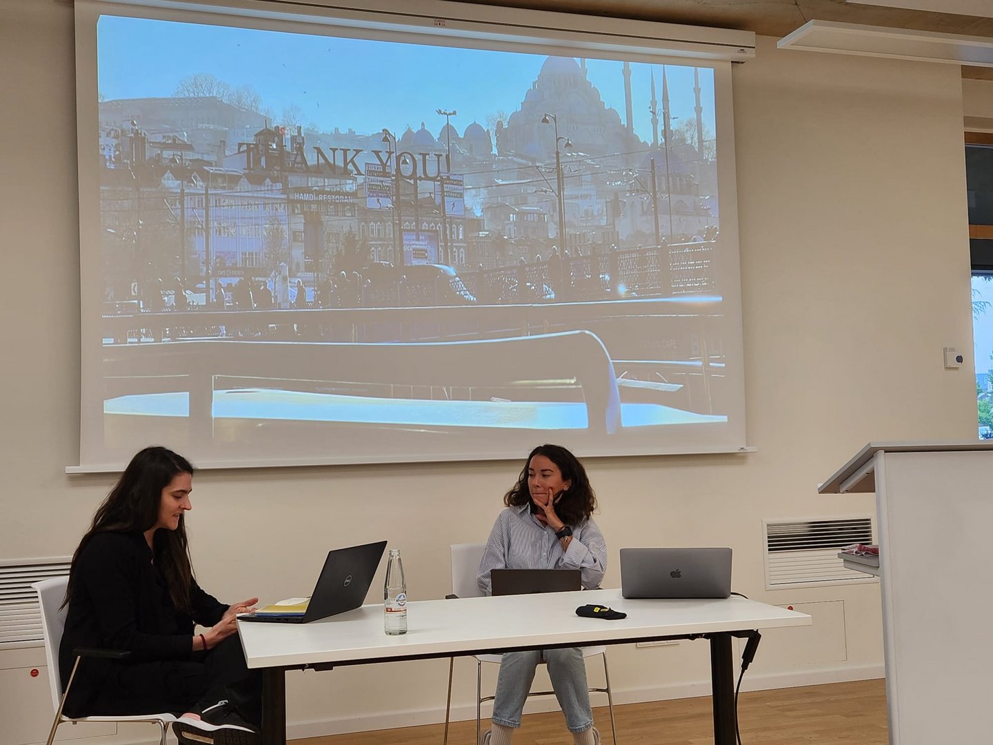 Two women with laptops, sitting in front of a presentation screen.